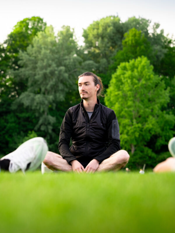 mentaltraining-tobias-waagwerke-meditation-training-anleitung Tobias von WAAGWERKE sitzt im Schneidersitz auf der Wiese und leitet eine Entspannungsübung zum Sport-Mentaltraining am Ende des Lauftrainings an.